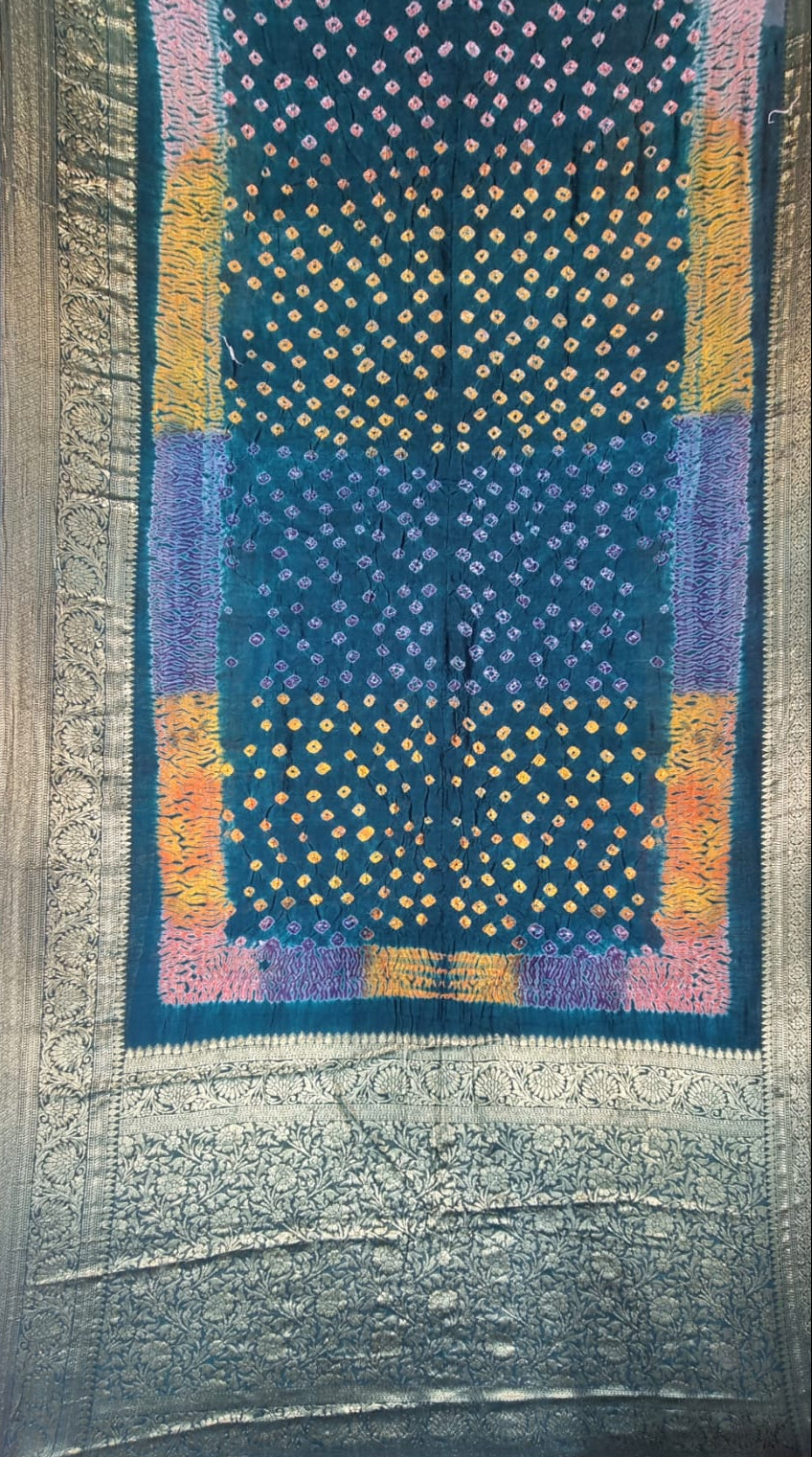 A folded patterned Art Silk Bandhani Saree with Shibori & Zari Border - Blue, featuring a dark teal body with yellow and pink bandhani-style dots, a purple border accent, and a wide metallic silver border.