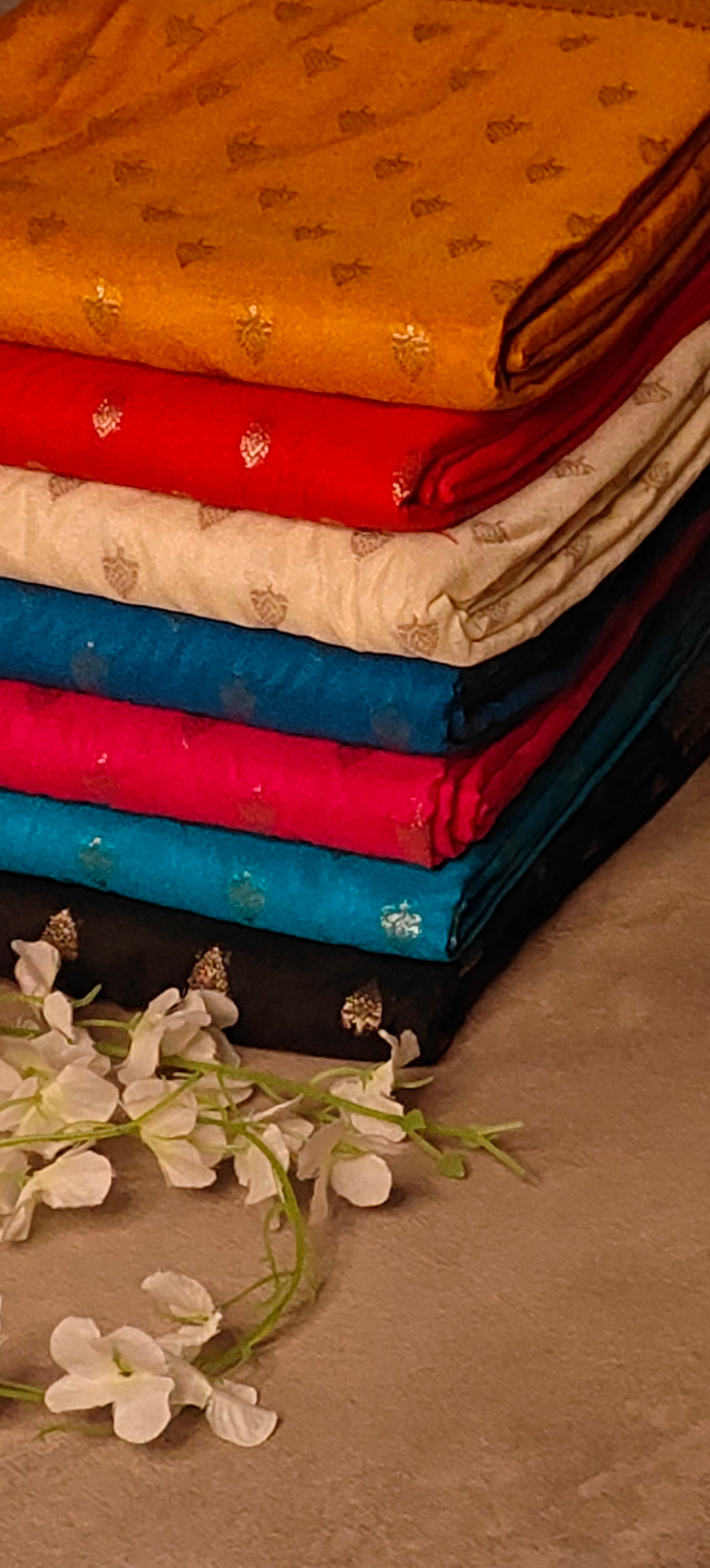 Blended Silk Saree with Khaddi Butti in Black, turqoise, magenta, teal, white, red, and in mustard yellow color stacked on beige background with flowers.