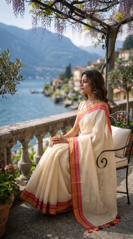 A side-profile shot of a woman relaxing on a wrought-iron chair on a scenic villa terrace balcony overlooking a blue lake and mountains, displaying the elegant drape of her white cotton saree