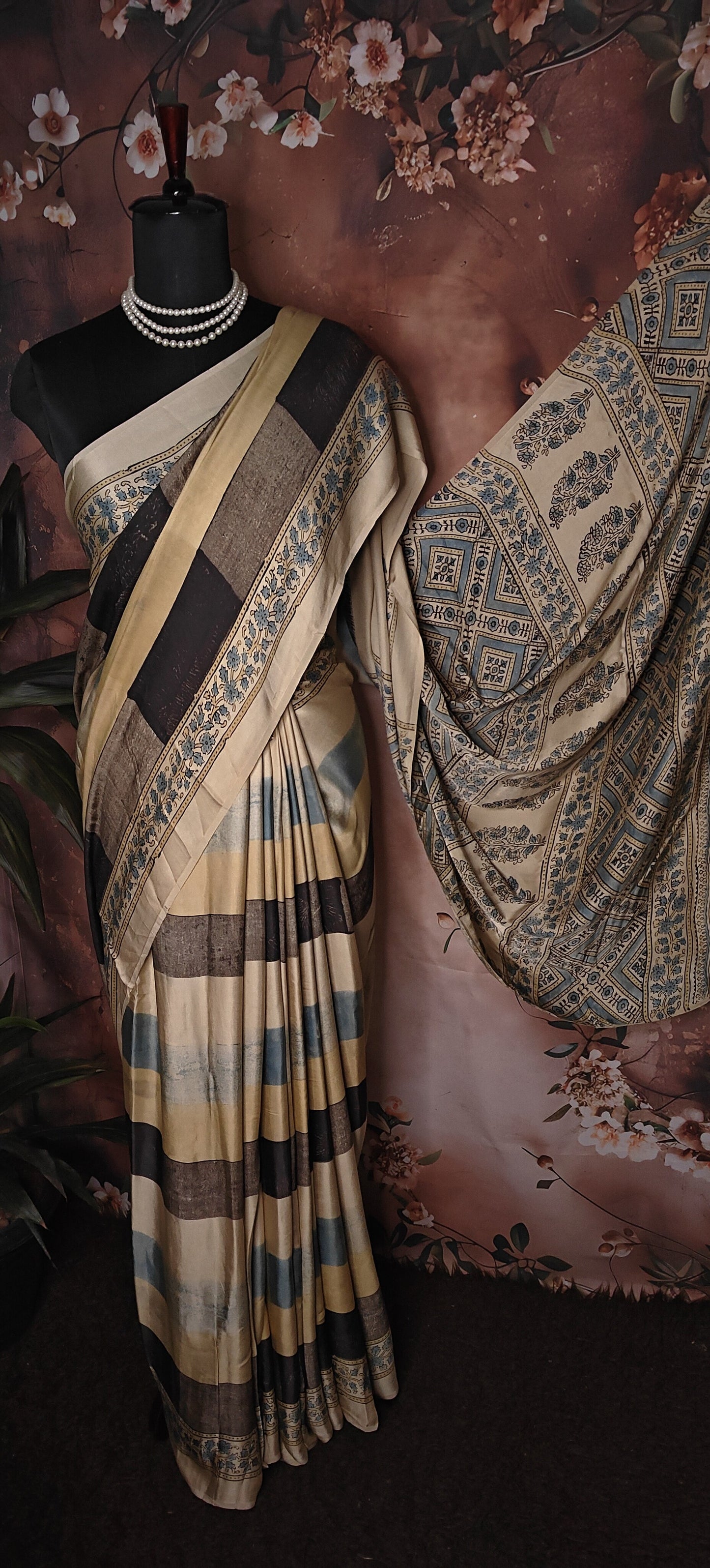 Ajrakh Modal Silk Saree – Beige, Black & Blue with Floral Border saree draped over a mannequin against a floral and fabric background