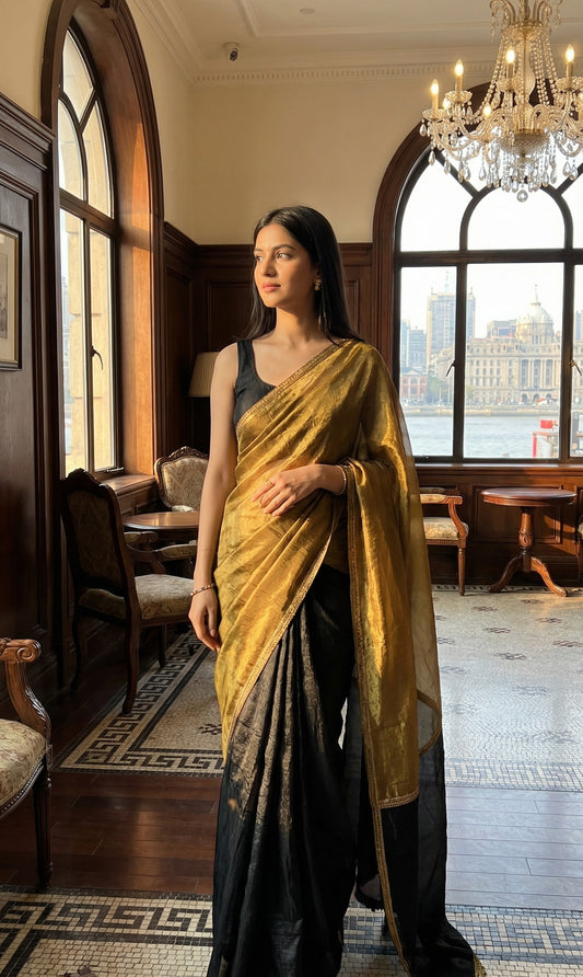 Woman in a antique golden Fendy silk saree with floral motifs and black contrast border, elegant festive wear, standing in a room with large windows and a chandelier.