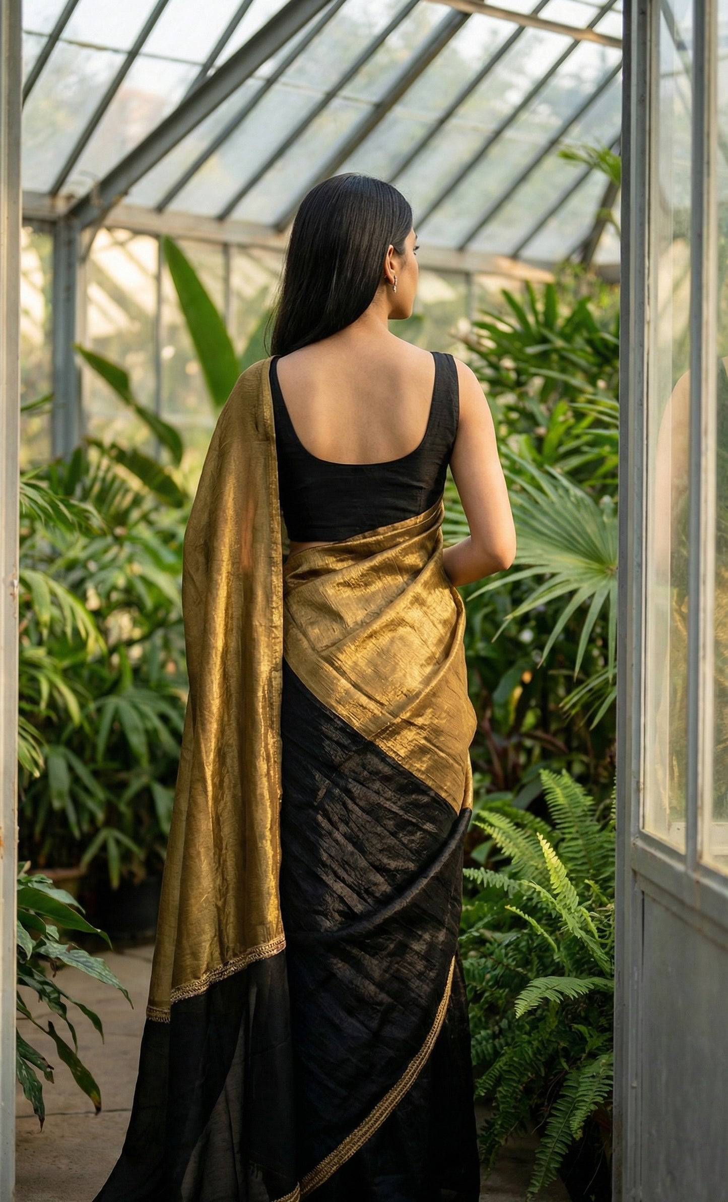 Woman in a antique golden Fendy silk saree with floral motifs and black contrast border, elegant festive wear, standing in a greenhouse filled with plants.