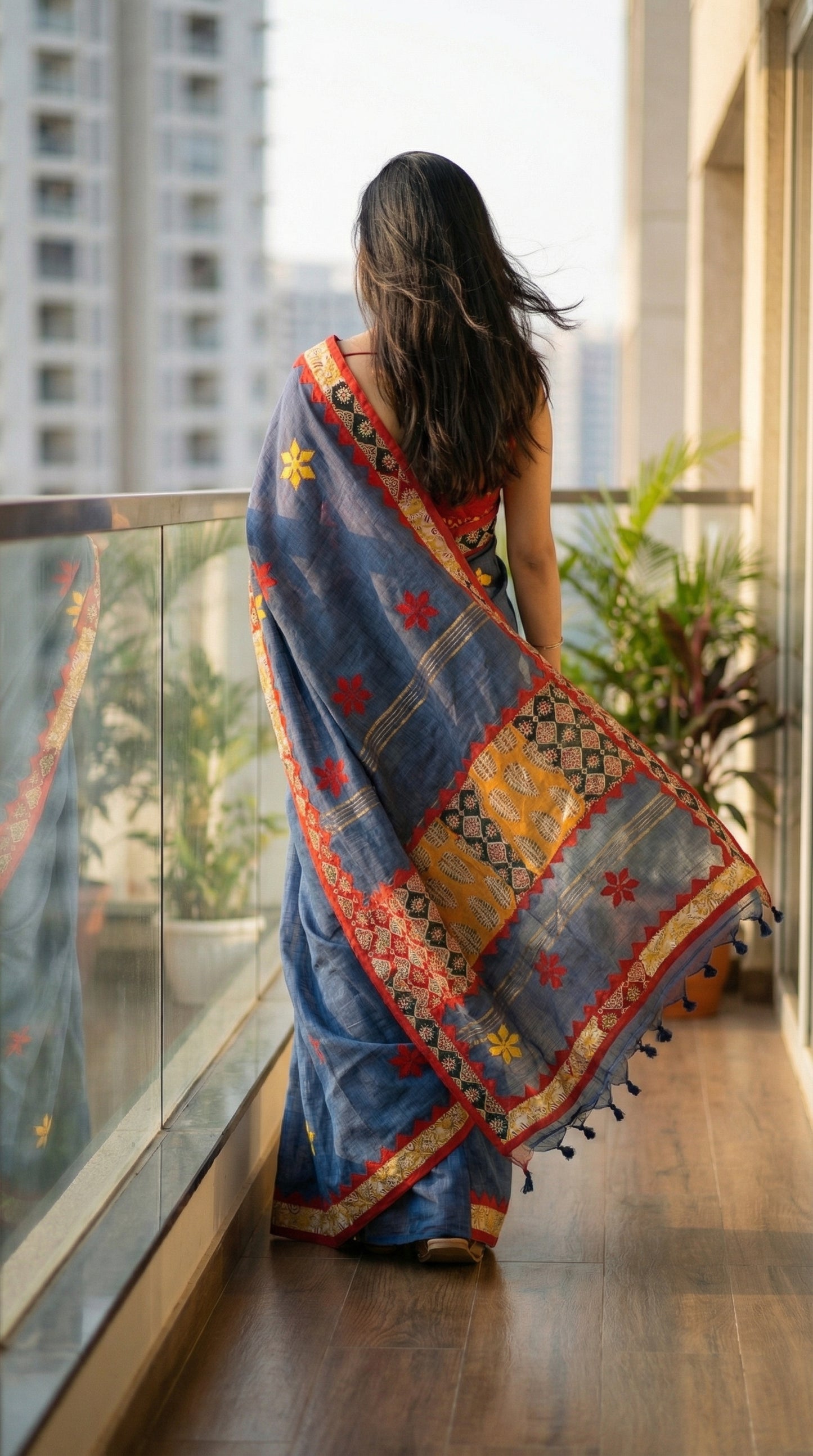 Woman in a grey linen cotton saree with traditional applique border, standing on a balcony with cityscape view.