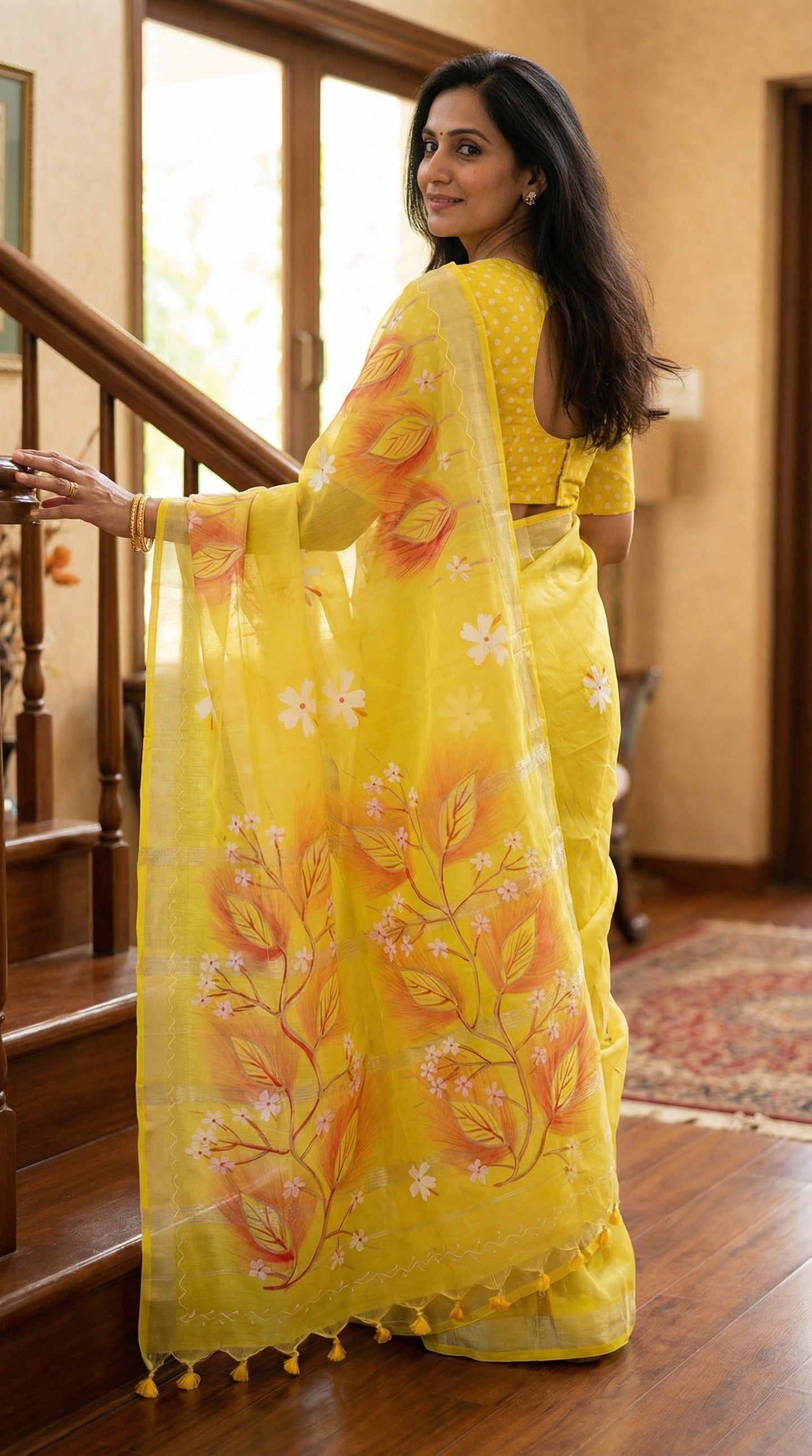 Woman in a yellow cotton blend saree with multicolor hand-painted floral and leaf motifs, lightweight artisanal saree, standing indoors.