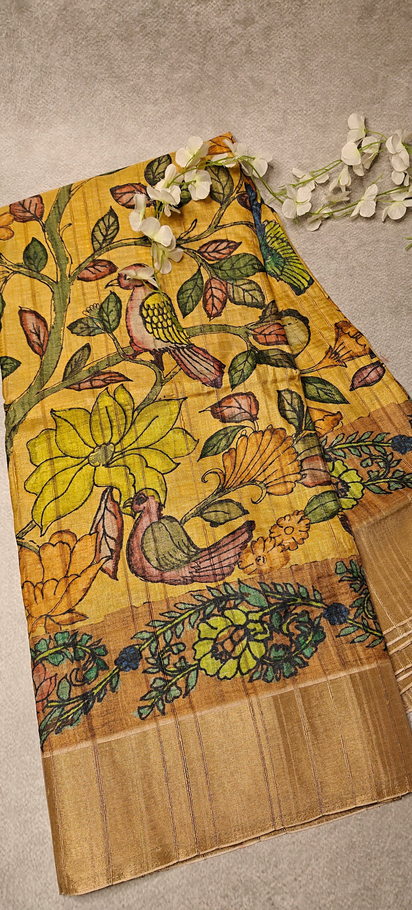 Yellow saree with Kalamkari-style bird and floral digital print and golden border lying flat and folded on a beige background with flowers.