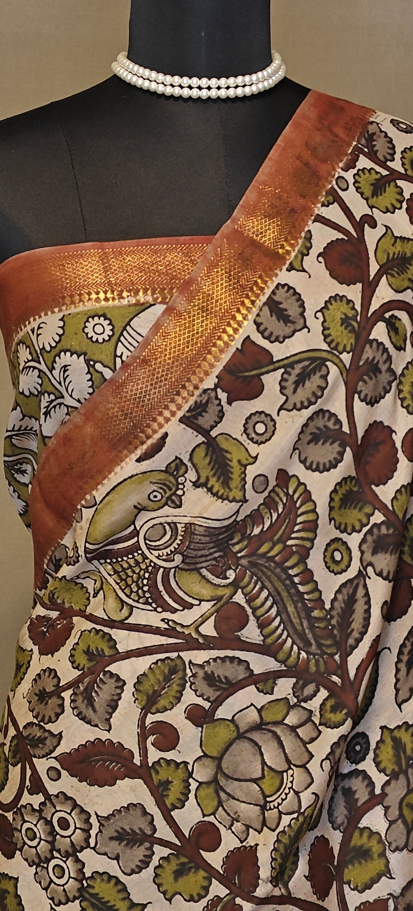 Brown silk saree with Kalamkari-inspired motifs and subtle golden border on a mannequin wearing a pearl necklace and blouse piece, against a neutral background.