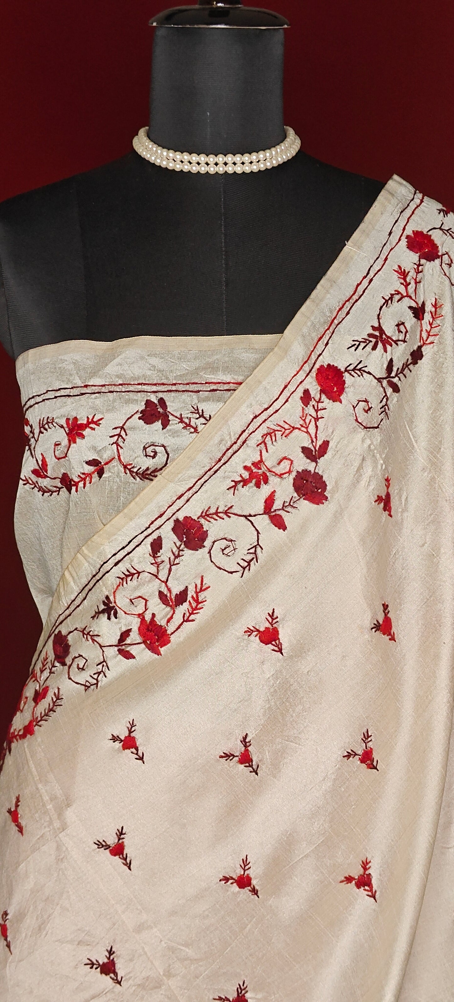 Handcrafted kantha stitched saree in cream with floral embroidery in maroon and red on a mannequin showcasing the blouse piece against a red background.