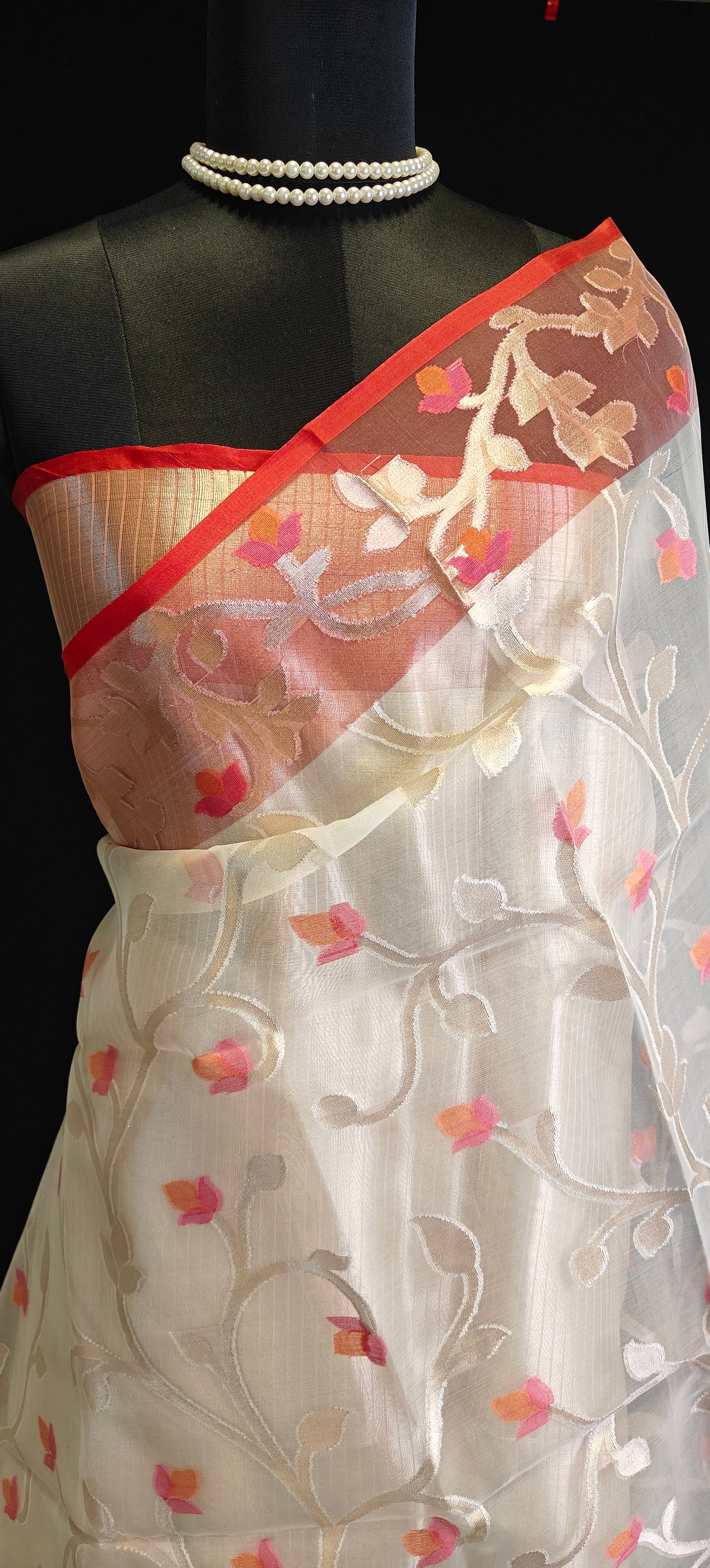 Cream muslin organza saree with red border and Jamdani floral embroidery, styled on mannequin with a flower vase and black background.