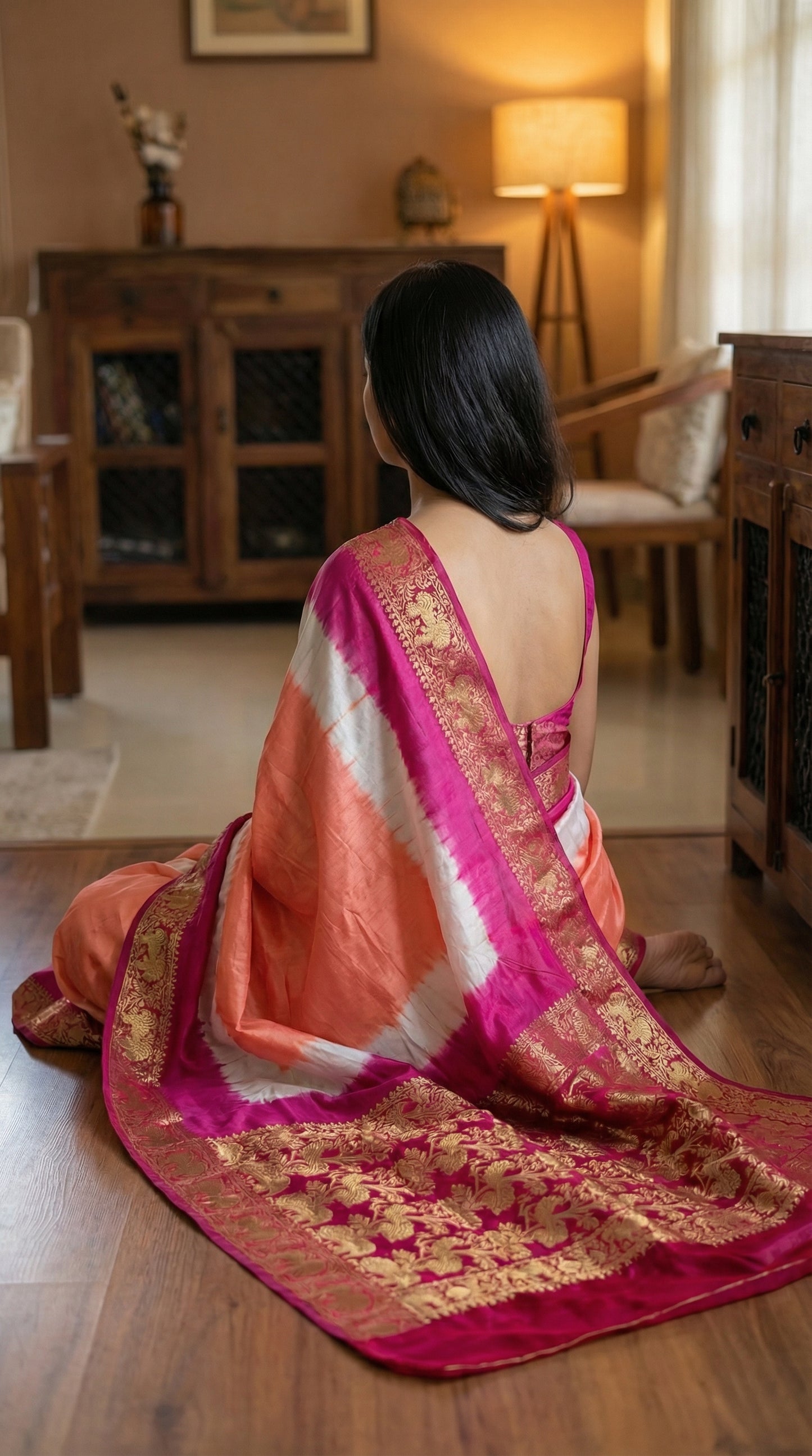 Woman in peach modal silk saree with pink Zari border and brocade pallu, sitting on the floor in a room with wooden furniture and a lamp.