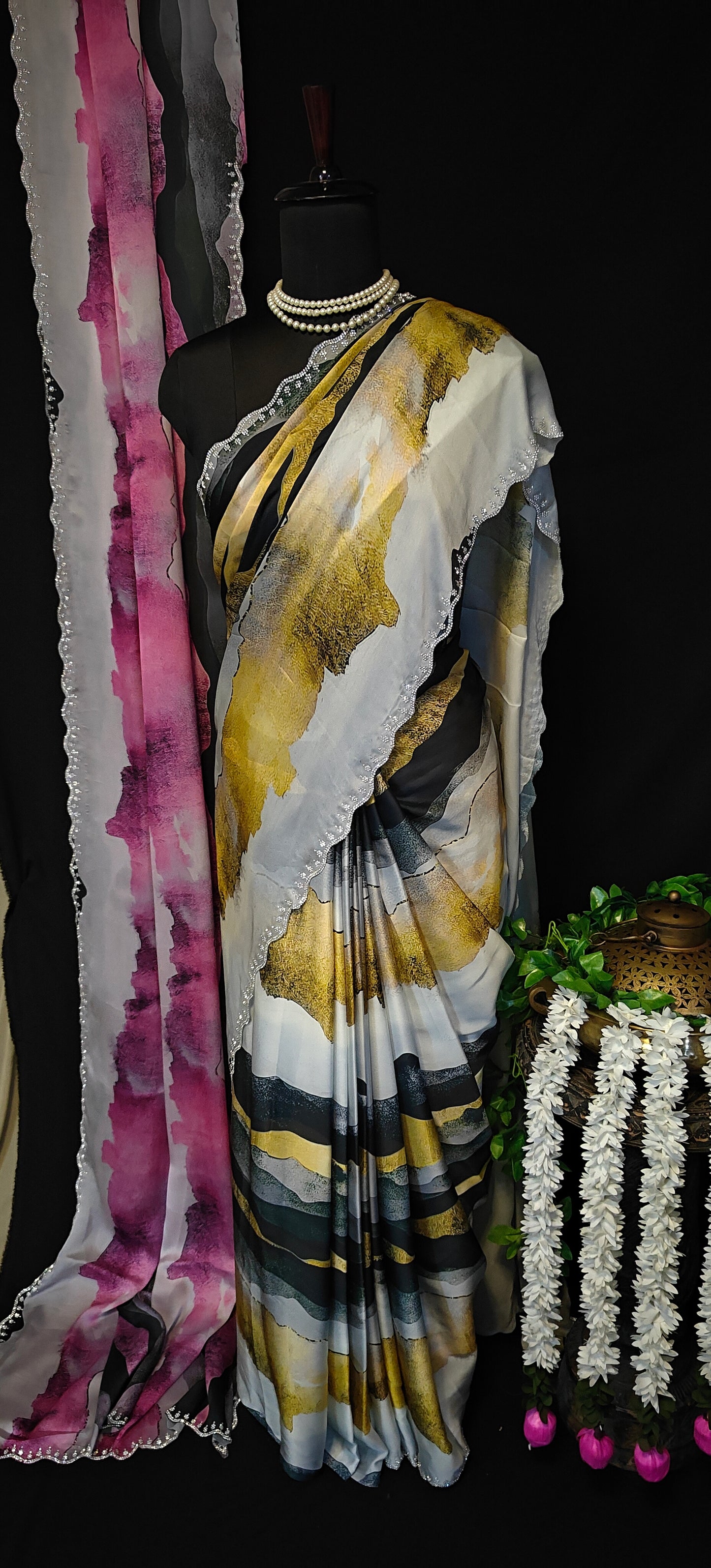 Pink satin saree with abstract print and scalloped silver border hanging next to a Yellow satin saree with abstract print and scalloped silver border plant with white flowers on a black background.