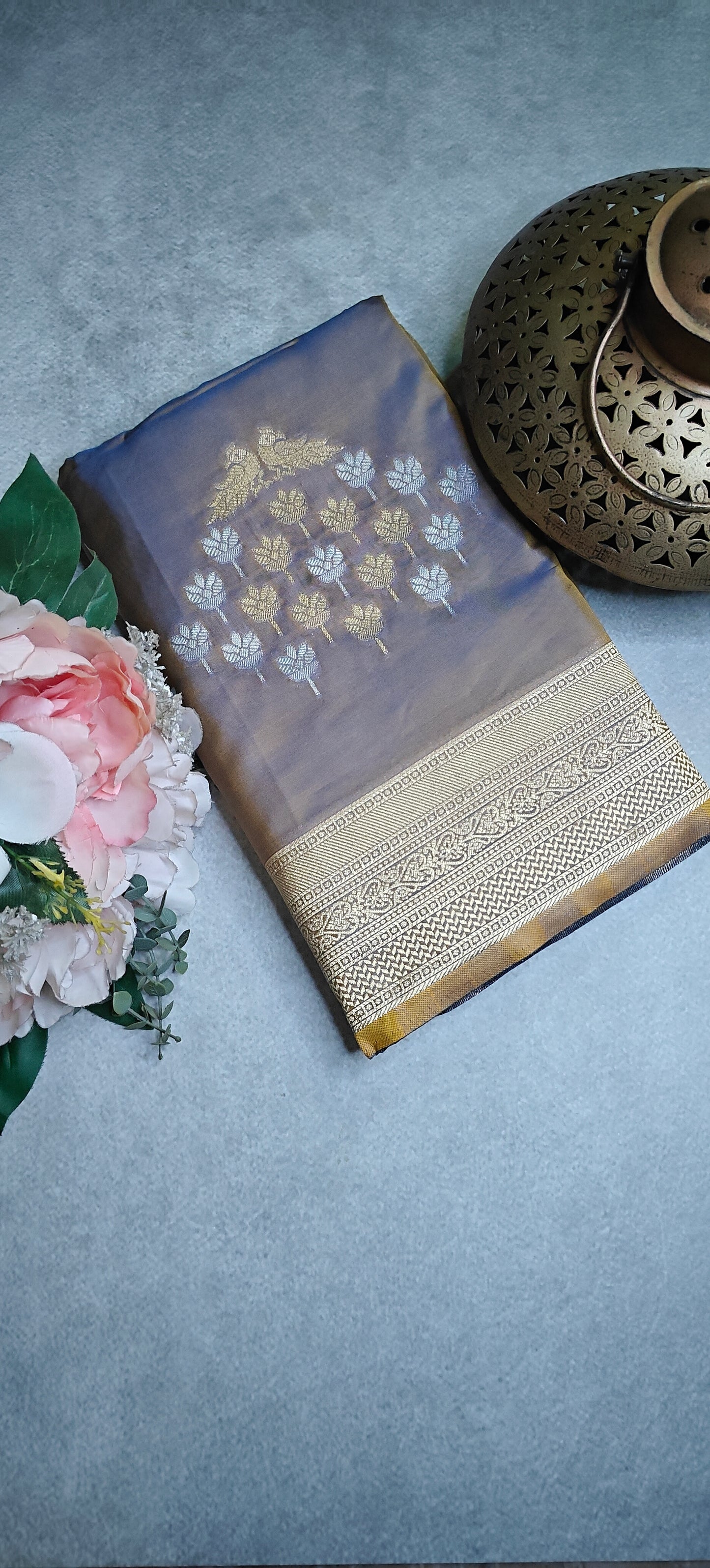 Grey soft silk saree with golden embroidery and floral zari border with flowers and a meal showpiece, lying flat on grey background.