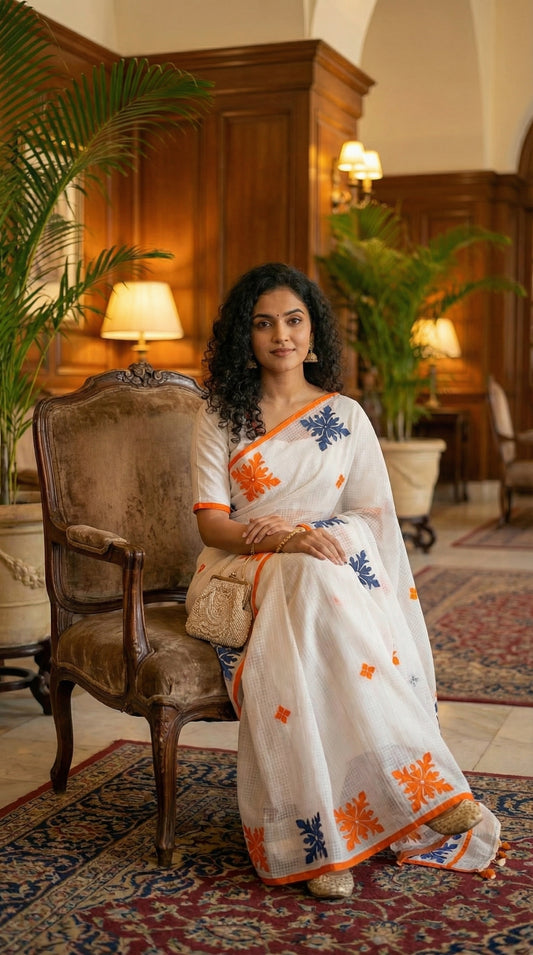 Woman in a white-orange-grey-applique-kota-saree, sitting on a decorative chair in an elegant room.