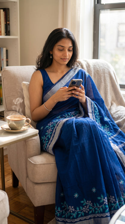 A woman sitting in an armchair using her phone with a steaming cup of tea nearby, wearing a royal blue cotton linen saree with light blue and white floral vine embroidery