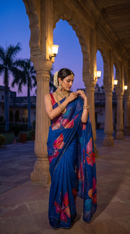 A model adorned in traditional heavy gold jewelry posing along a lit palace colonnade at twilight, highlighting the premium shine and detailed cutdana work on her printed blue saree