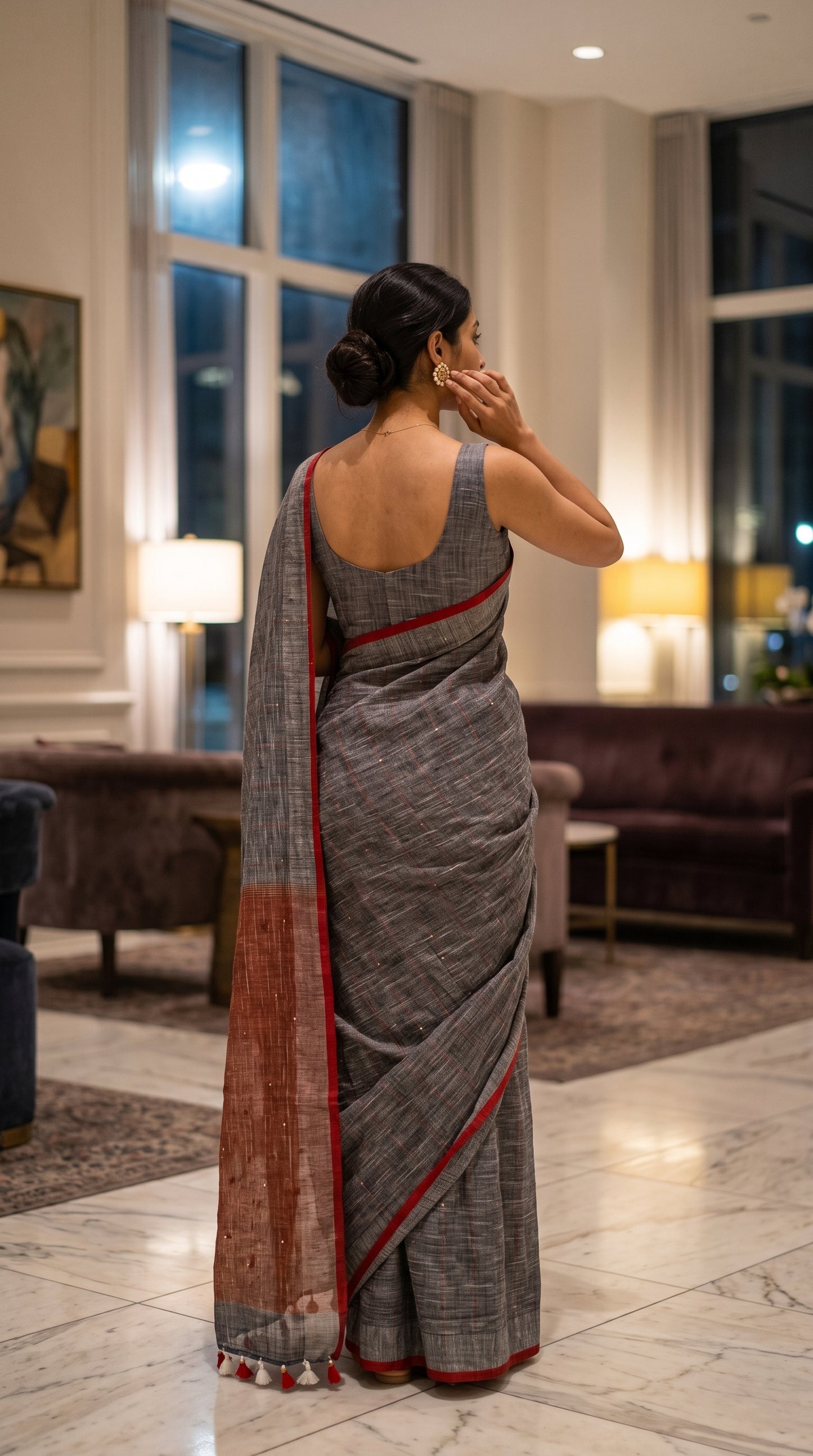 Back view of a woman standing in a boutique hotel lobby adjusting her earring, highlighting the elegant fall, seamless blouse fit, and bright red border trim of her charcoal grey saree