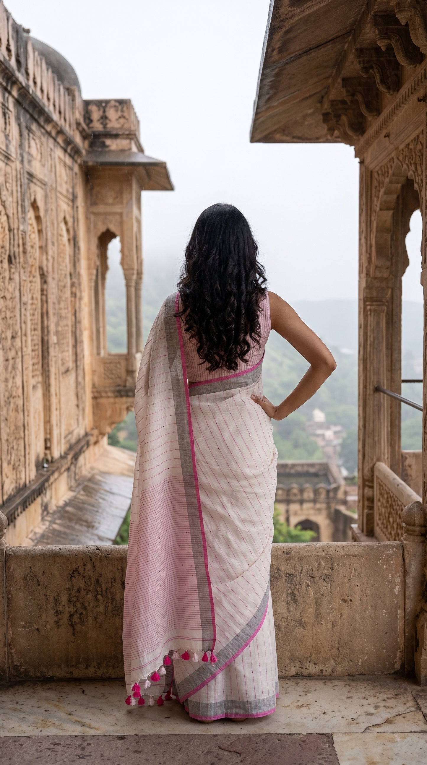 Back view of a woman looking out from a stone palace balcony over a misty valley, highlighting the striped pallu and pink hanging tassels of a white khadi linen saree
