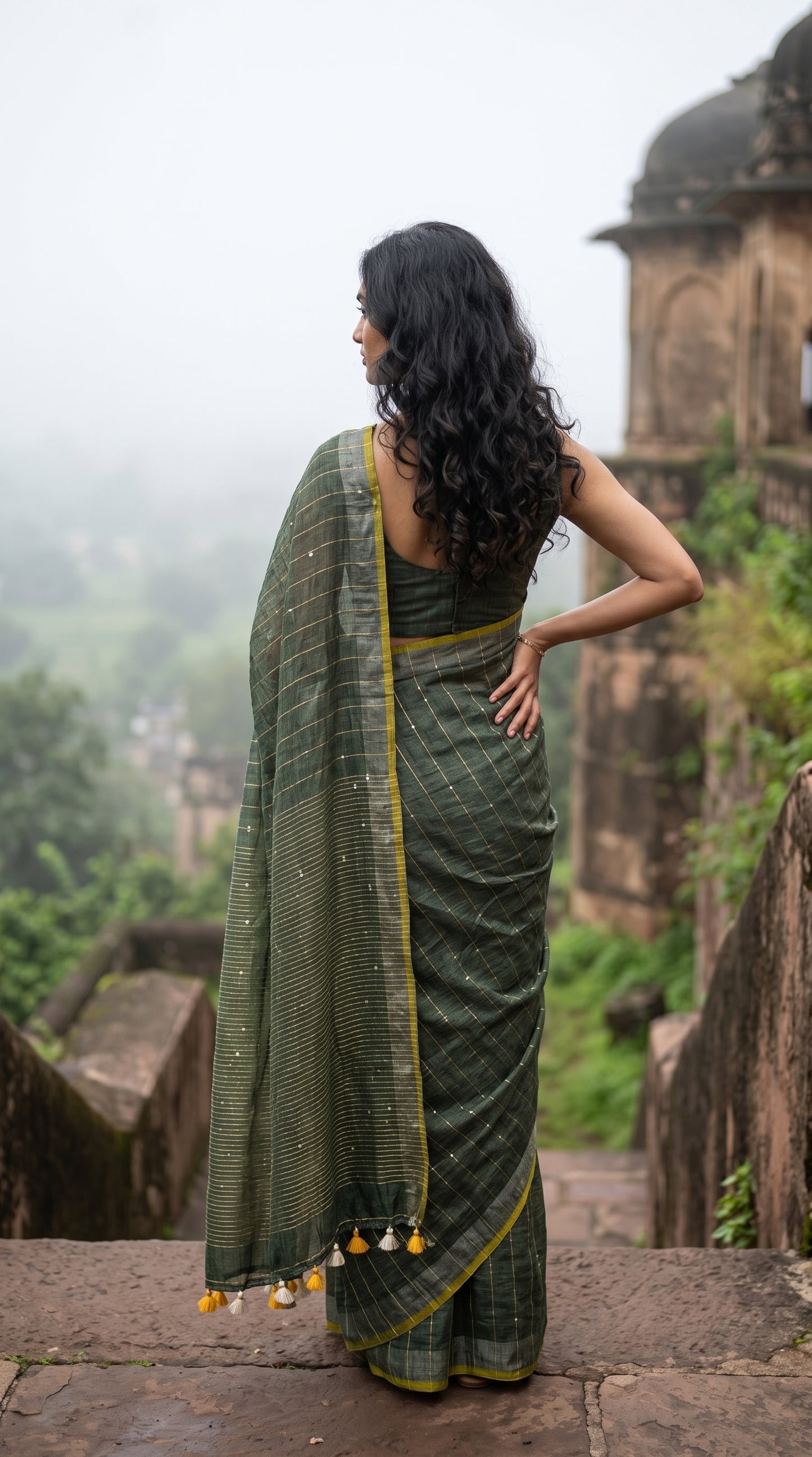 Back view of a woman standing on stone steps overlooking a misty green valley during the monsoon, displaying the vertical gold stripes, silver border, and yellow tassels of an olive green khadi linen saree