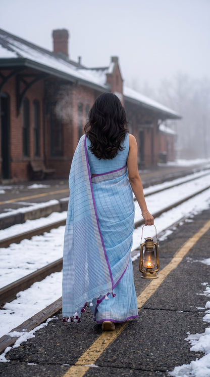 Back view of a woman carrying a vintage lantern down a snowy railway platform, highlighting the horizontal pinstripe pattern on the pallu and dark purple tassels of her sky blue khadi linen saree