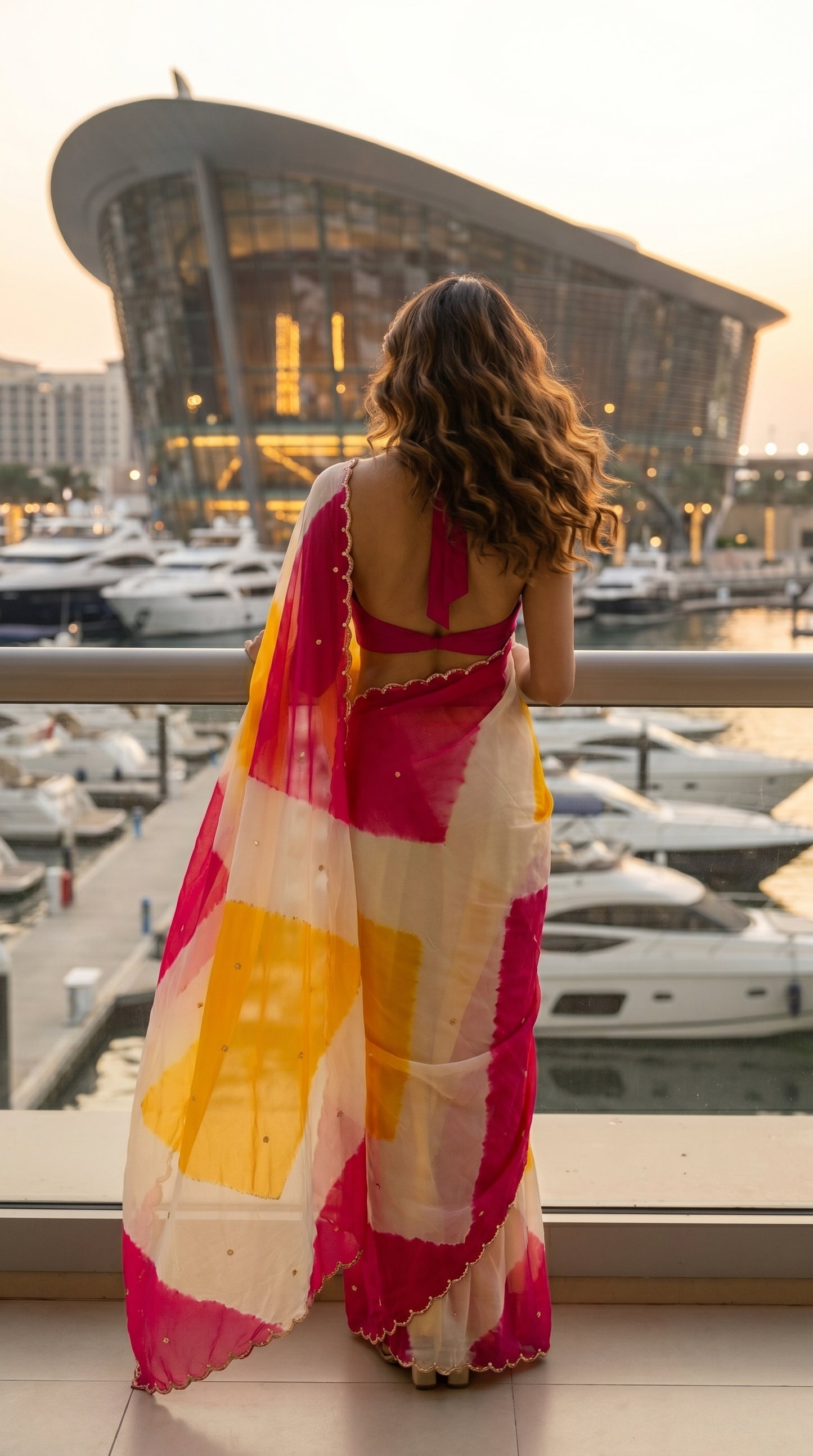 Back view of a woman looking out over a marina, highlighting the graceful movement and scalloped pallu drape of an ivory and pink chiffon saree