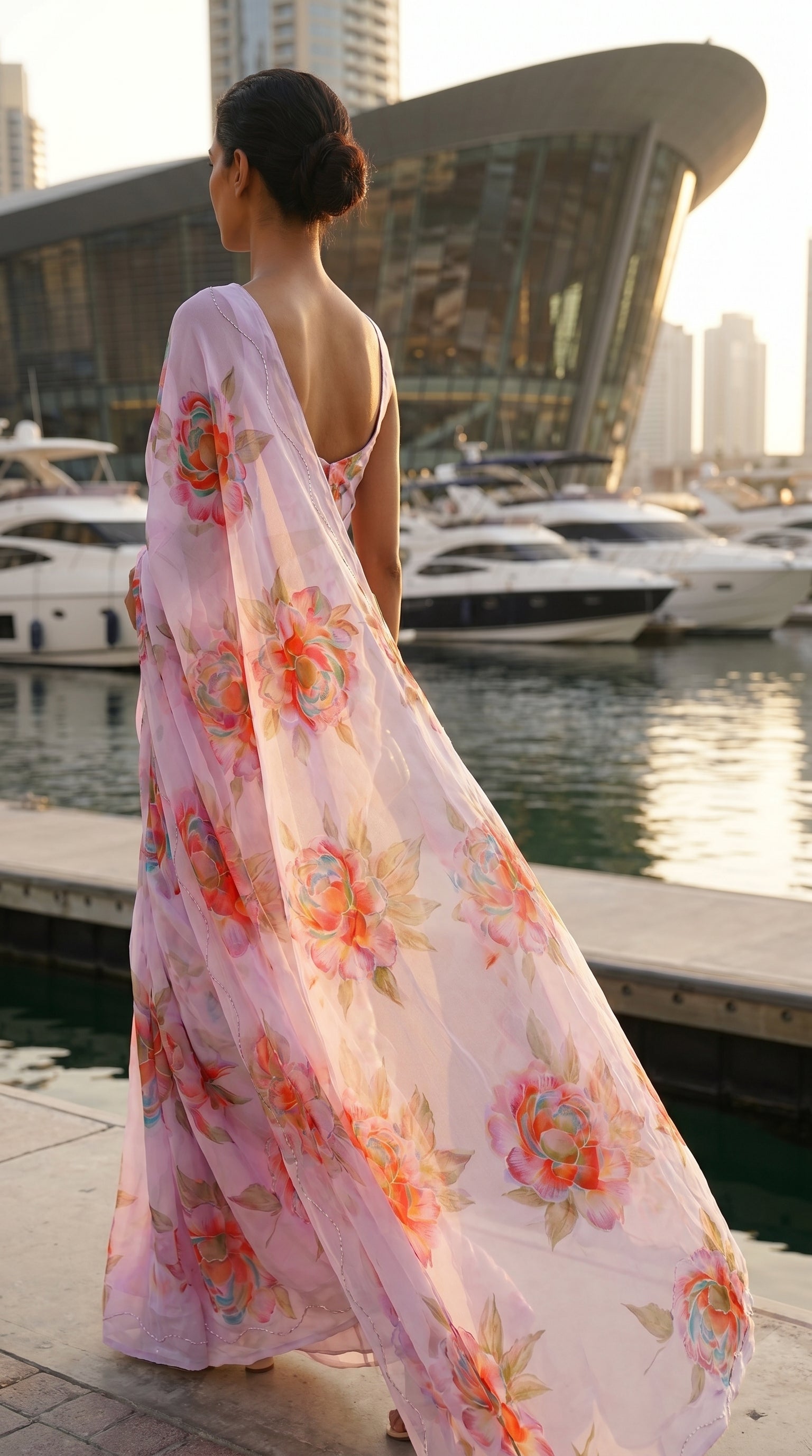 Back view of a woman standing on a marina deck at dusk, showcasing the long, sweeping trail and detailed cut work borders of a light lavender floral chiffon pallu
