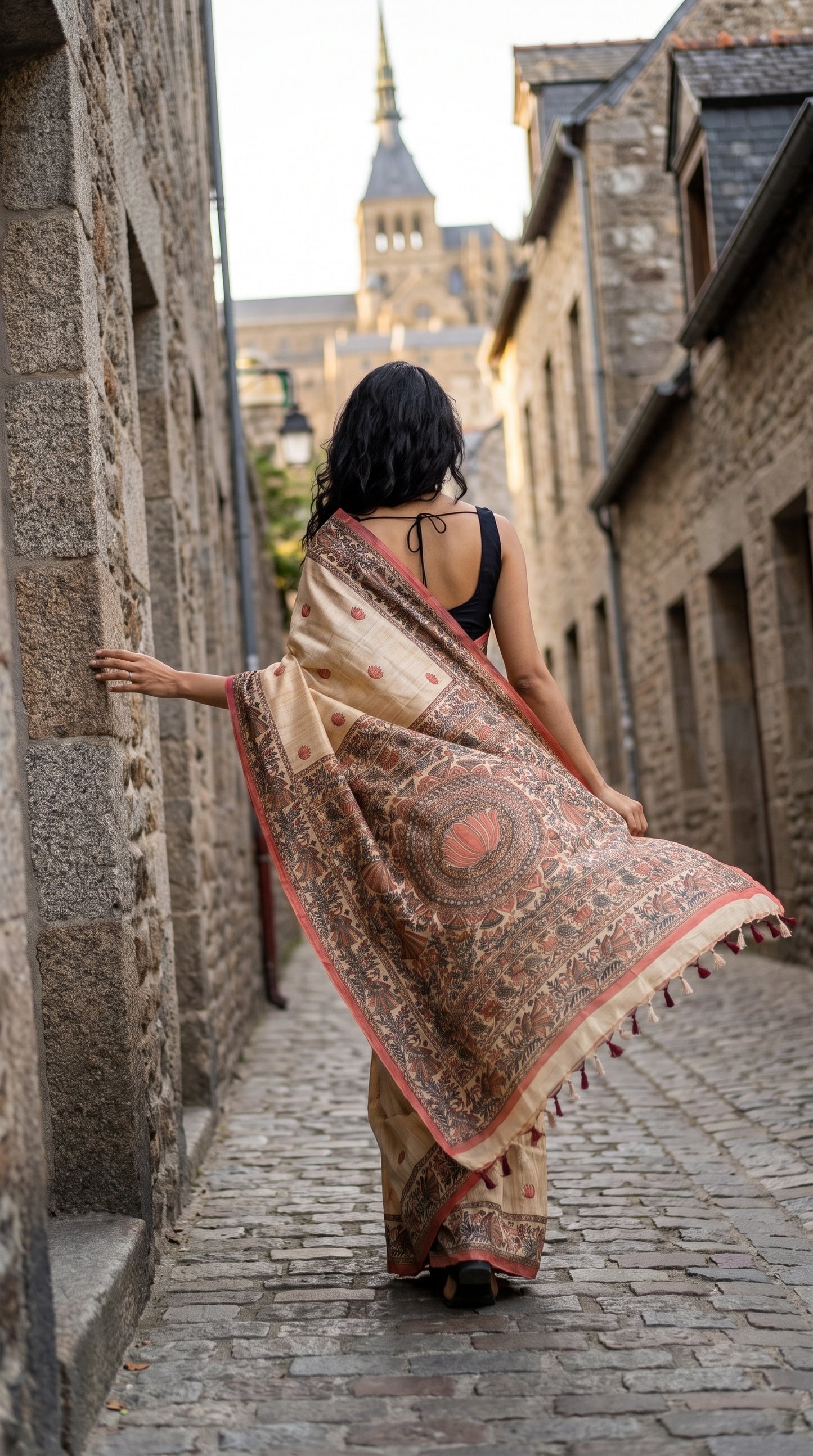 Back view of a woman walking down a narrow stone street, showcasing a beige Tussar silk saree with a heavily detailed light-red Madhubani printed pallu and edge tassels