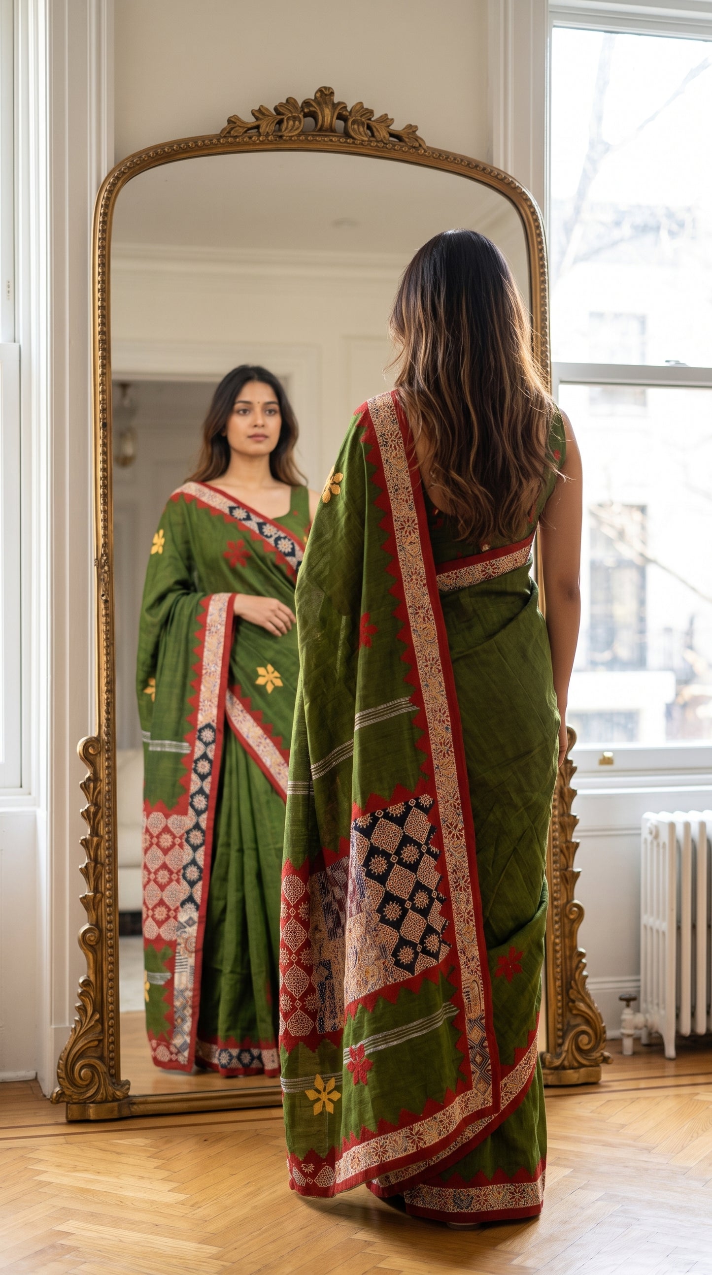A woman standing with her back to the camera looking into a large gilded full-length mirror, showing the intricate red and black patchwork panels on her olive green saree