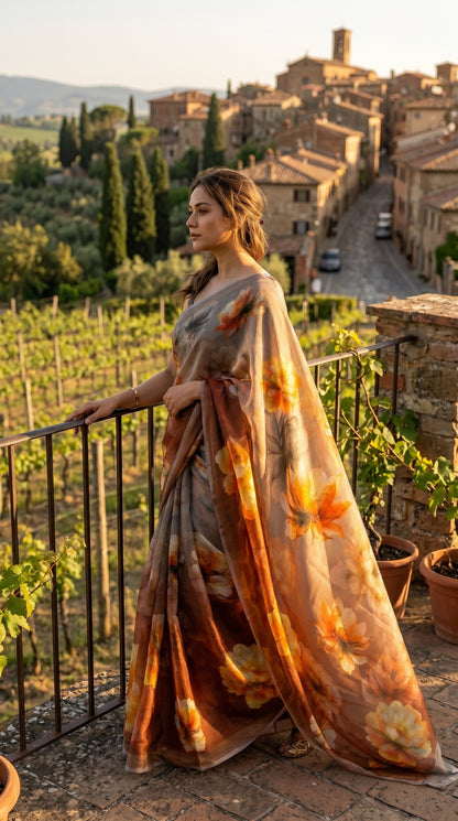 Model leaning against a terrace railing overlooking a sun-drenched vineyard, showcasing the rich golden and terracotta peony motifs on her brown chiffon pallu