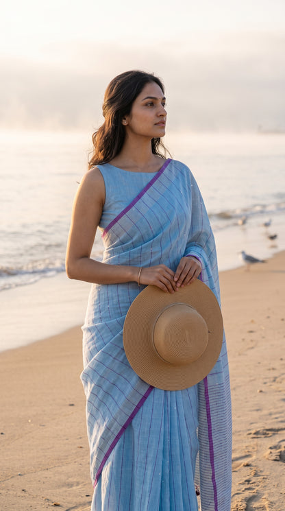 A portrait of a woman holding a straw hat while standing on a beach shoreline at sunrise, displaying the refreshing sky blue color and vertical purple stripes of her khadi linen saree