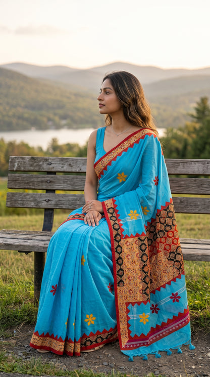 Model seated on a rustic wooden bench looking out at green rolling hills and a lakeside view, beautifully spreading out the heavy printed patchwork pallu and fringe tassels of a sky blue saree