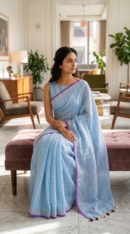 Model seated on a plush pink velvet bench inside a bright boutique hotel lobby, showcasing the elegant pleats and purple-and-grey dual-tone borders of her sky blue saree
