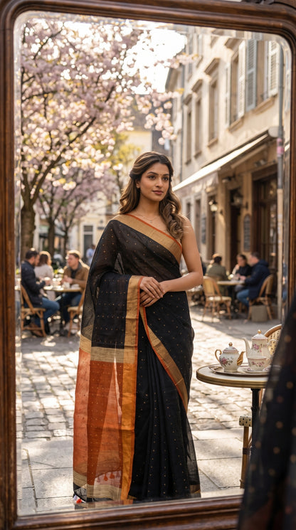 Model sitting at a Parisian-style street café table looking into a handheld mirror, highlighting the drape of her black linen saree and the translucent contrast orange pallu