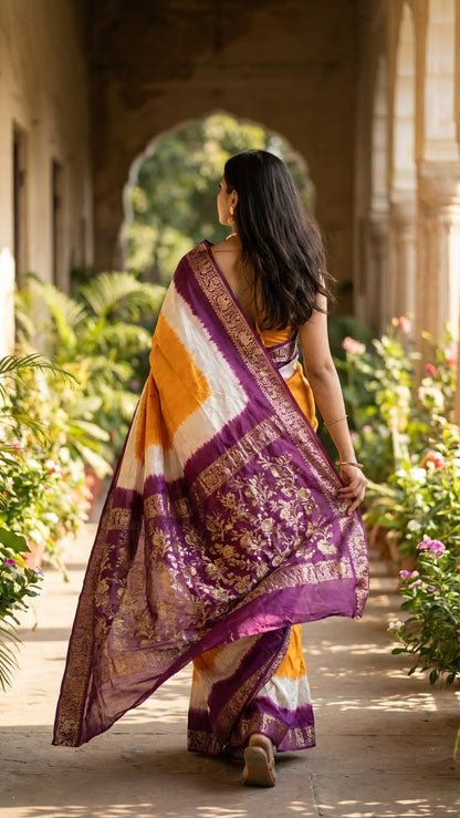 Woman in a golden-orange modal silk saree with purple zari border– designer saree for festive events walking through a garden with architectural elements.