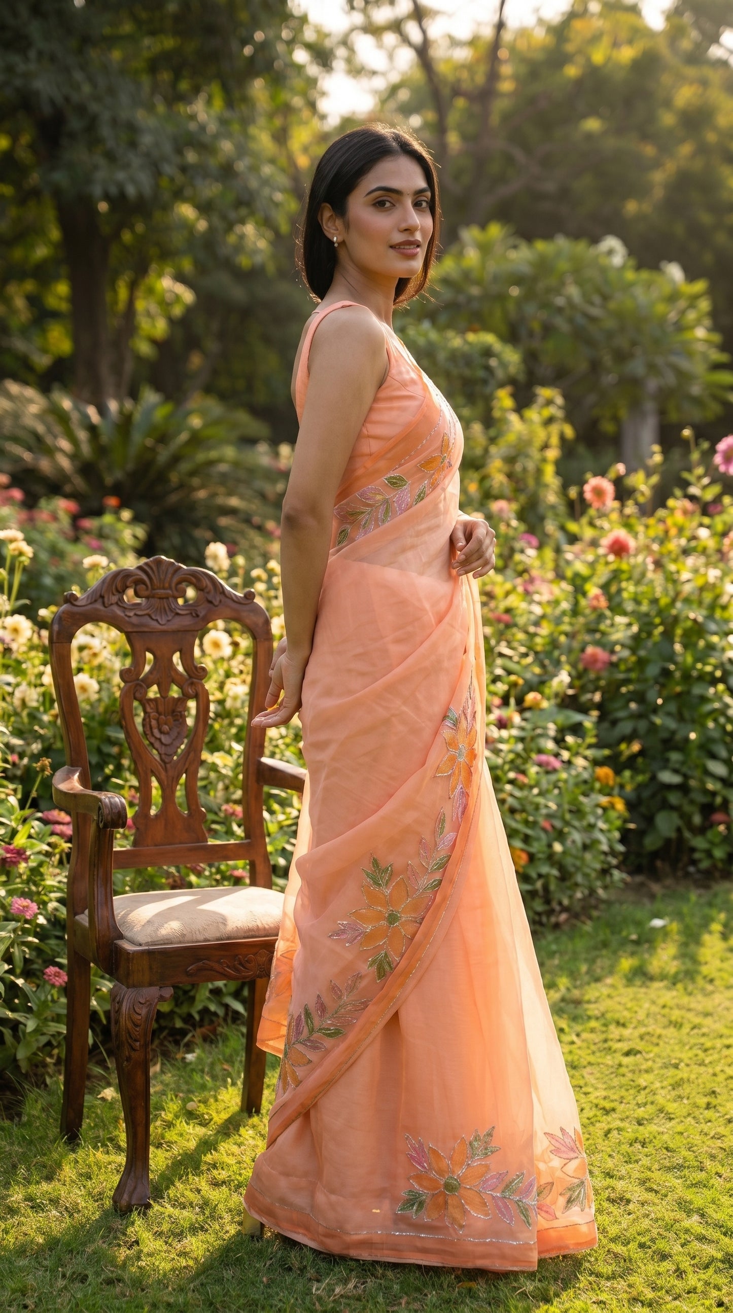Full-length image of a woman posing on a lawn next to a classic wooden chair, displaying the Super-soft texture, delicate translucency, and trailing handworked pallu of her peach pure chiffon saree