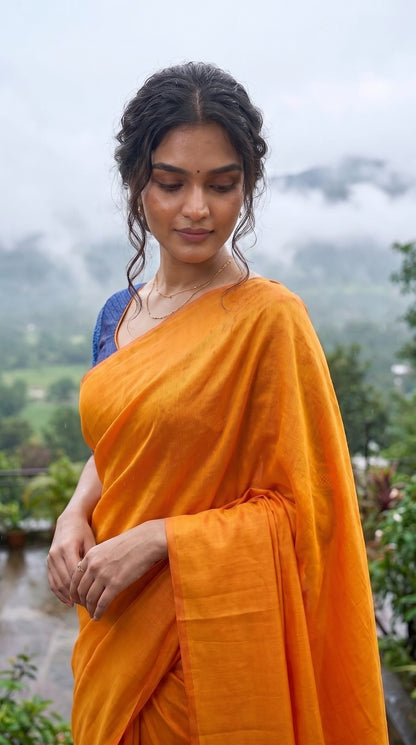 A portrait of a woman looking down thoughtfully against a backdrop of misty mountains on a monsoon morning, showcasing the rich fabric texture and soft metallic glaze of her orange tissue cotton saree
