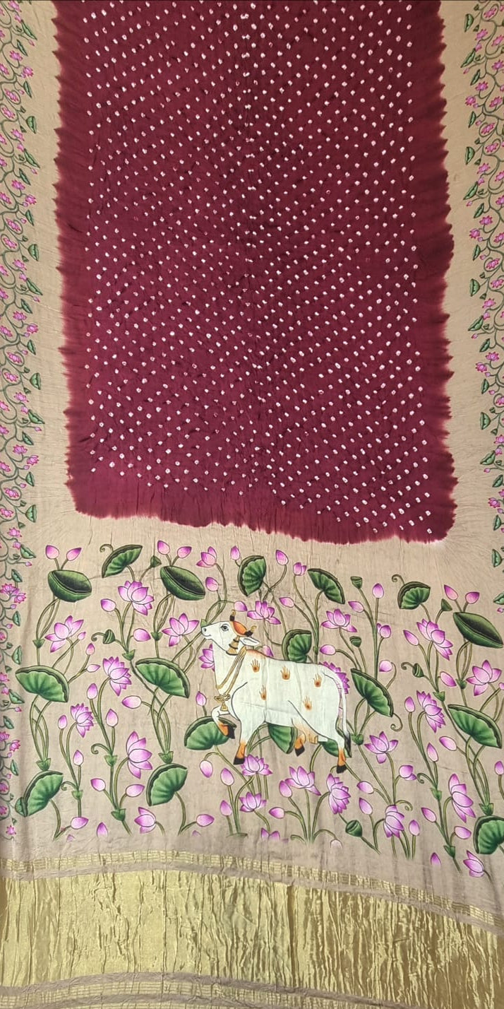 A Modal Silk with Bandhani Pichhwai Print Border - Maroon saree displayed flat, featuring a deep maroon red body with white dot work, a beige border with pink floral embroidery, and a gold zari border. The lower section is decorated with a hand-embroidered cow surrounded by pink flowers and green leaves.