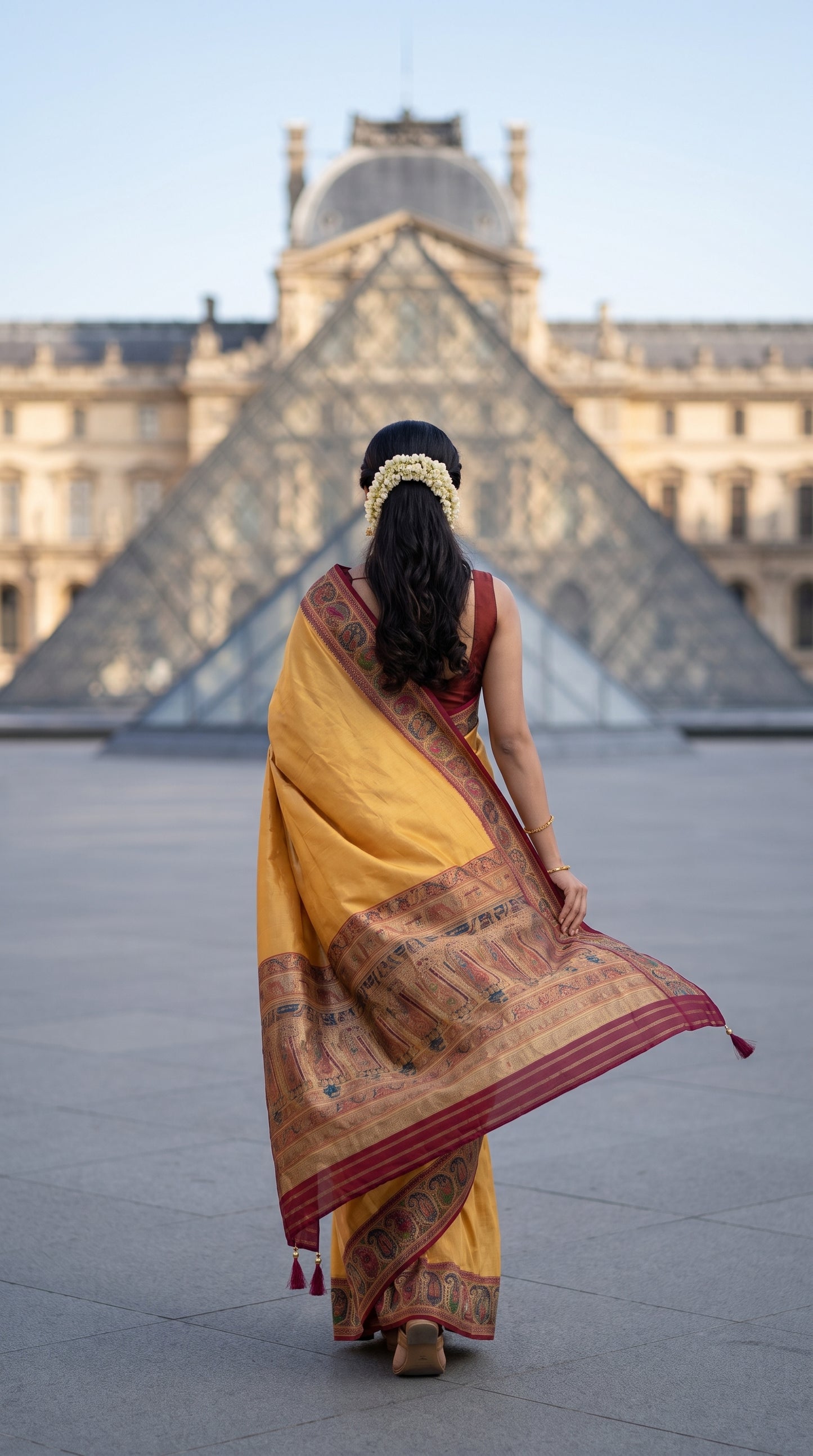 A rear view of a woman with a fresh gajra hair decoration walking toward the symmetrical glass pyramid structure at the Louvre Museum, showcasing the floating pallu and rich multicolored jamawar borders of her beige Kani silk saree