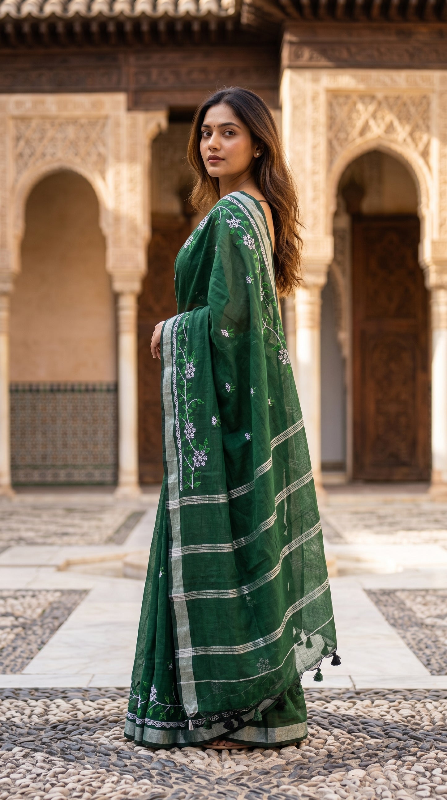 Side profile view of a woman standing in a stone courtyard with historic archways, showcasing the structured pallu with its silver Zari striped pattern and hanging edge tassels