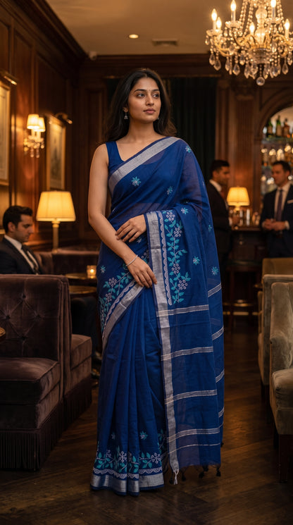 Model standing gracefully inside a dimly lit wood-paneled lounge beneath a chandelier, showcasing the silver Zari border and intricate floral embroidery of a blue cotton linen saree