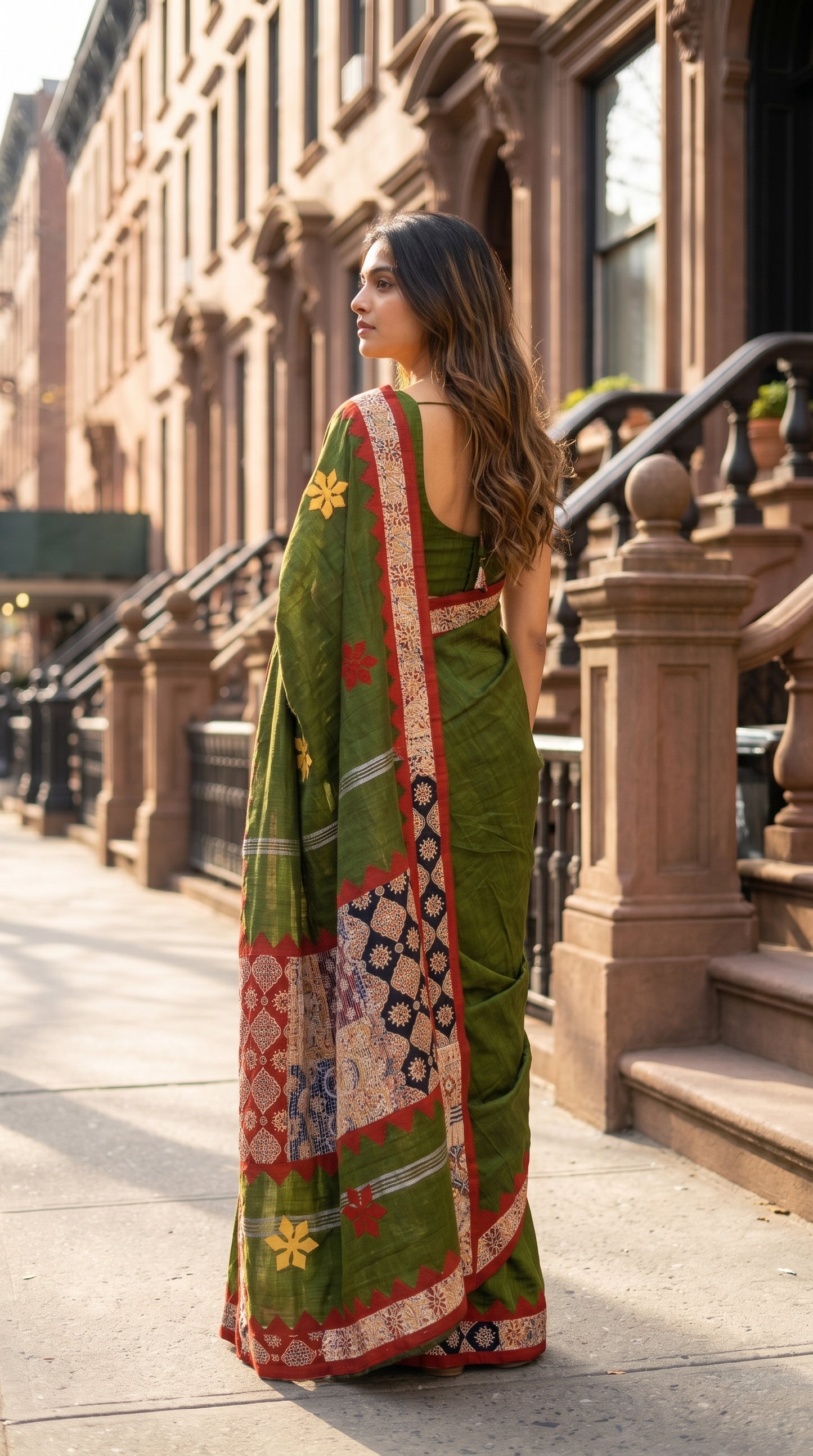 Three-quarter back view of a woman walking past brownstone buildings, showcasing the detailed multi-patterned geometric patchwork pallu of an olive green saree