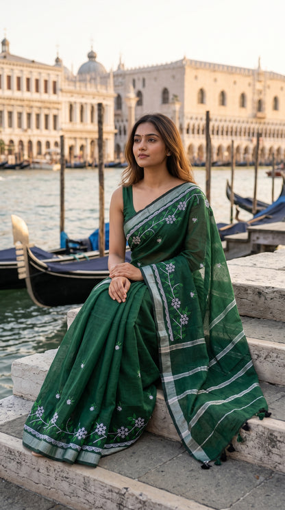 A model sitting on stone steps next to a Venice canal with gondolas and historic palaces in the background, draped in a forest green cotton linen saree with white floral vine embroidery
