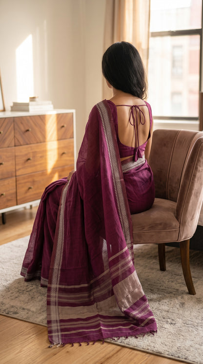 Back angle view of a woman sitting on a brown armchair, showing the elegant tie-back blouse and the flowing striped pallu of a magenta mul cotton saree