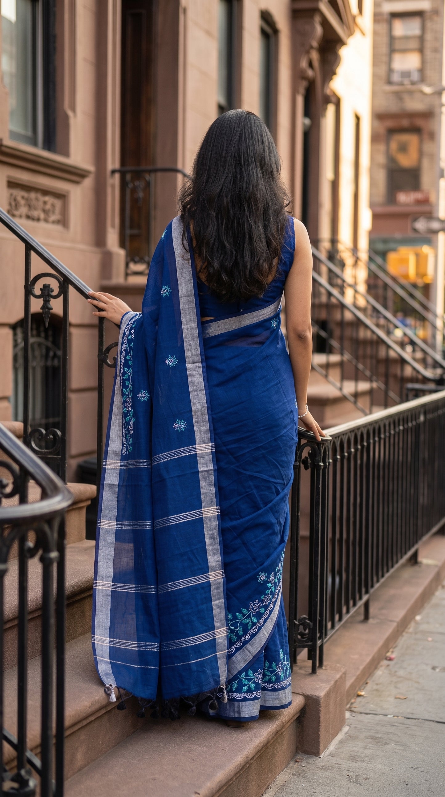 Back view of a woman walking up brownstone steps holding the iron railing, displaying the elegant striped silver Zari pallu and hanging tassels of her blue linen blend saree