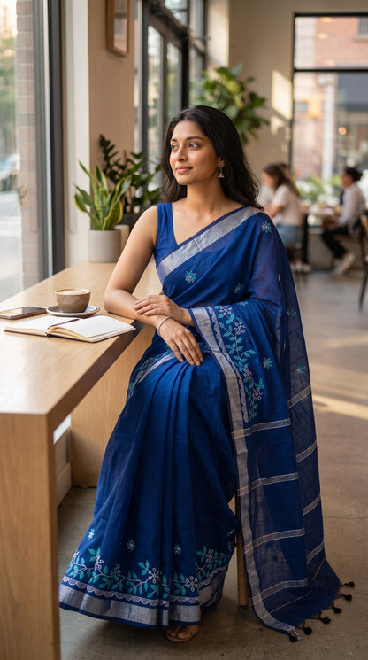 A model sitting by a cafe counter looking out the window, highlighting the crisp drape, silver highlights, and detailed floral border artwork on her royal blue saree