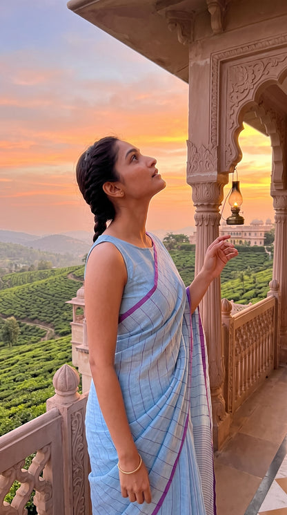 A model looking out from a carved stone palace balcony overlooking rolling green tea hills at sunset, displaying the neat silhouette of her sky blue sequined tissue saree