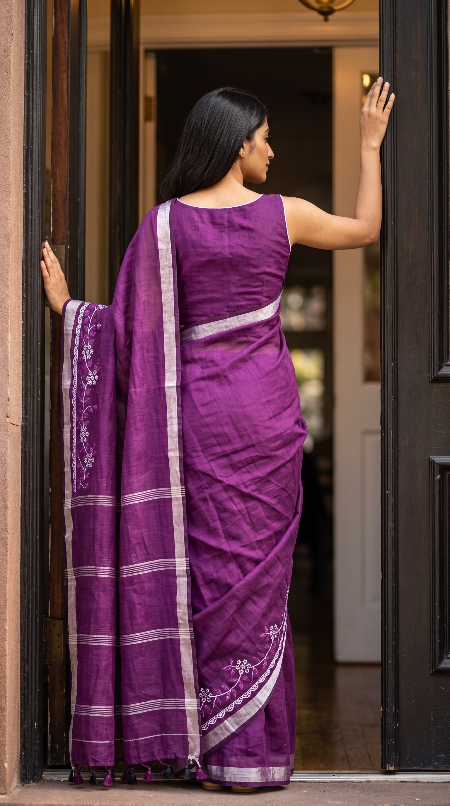 Back view of a woman standing in a doorway with her hand on the frame, displaying the clean lines of the blouse and the striped silver Zari pallu of a purple saree