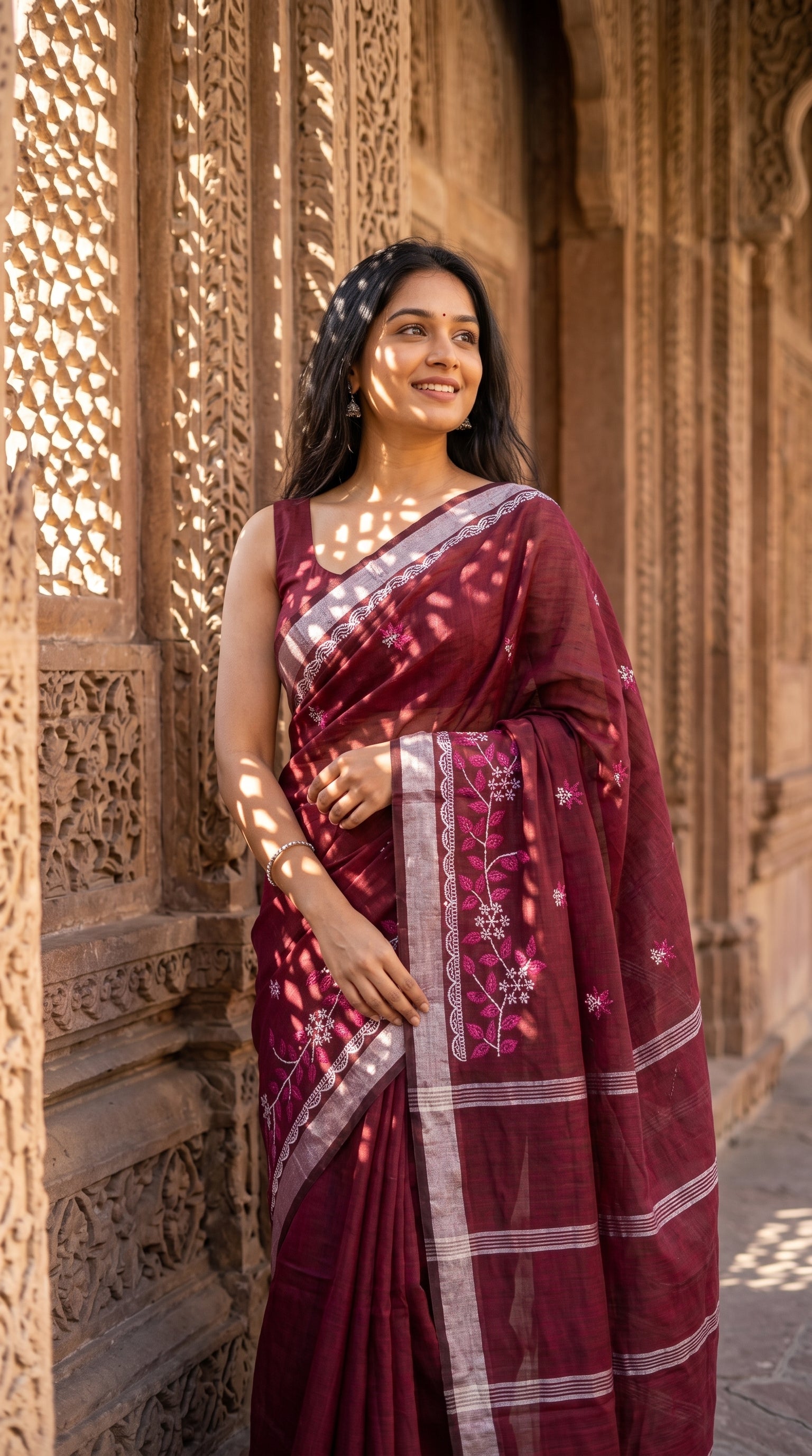 A woman standing beside an intricately carved stone monument wall with geometric shadow patterns, highlighting the delicate floral motifs on her maroon saree
