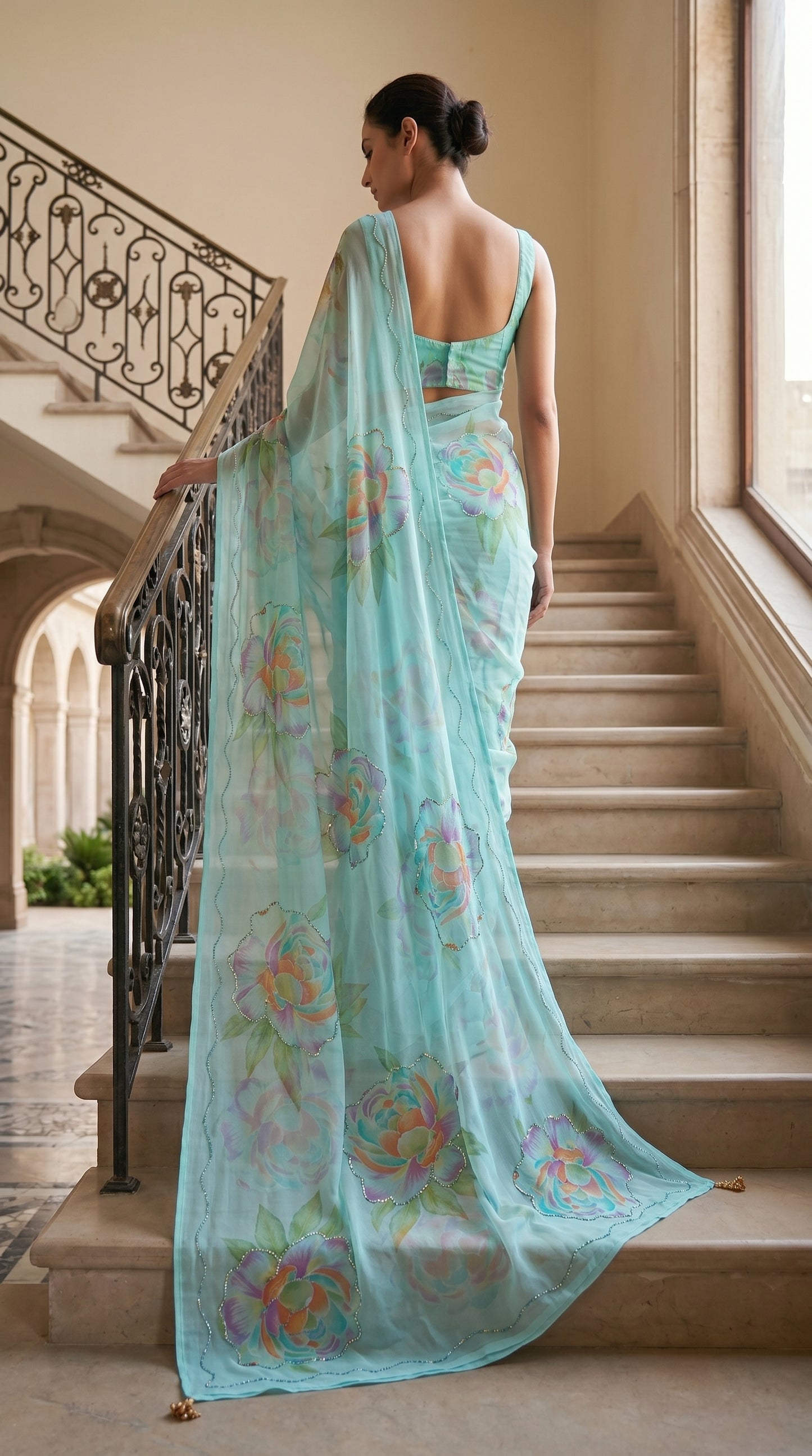 Full-length back view of a model descending marble stairs, displaying the sweeping pallu trail and scalloped edge cut work of an aqua blue floral chiffon saree