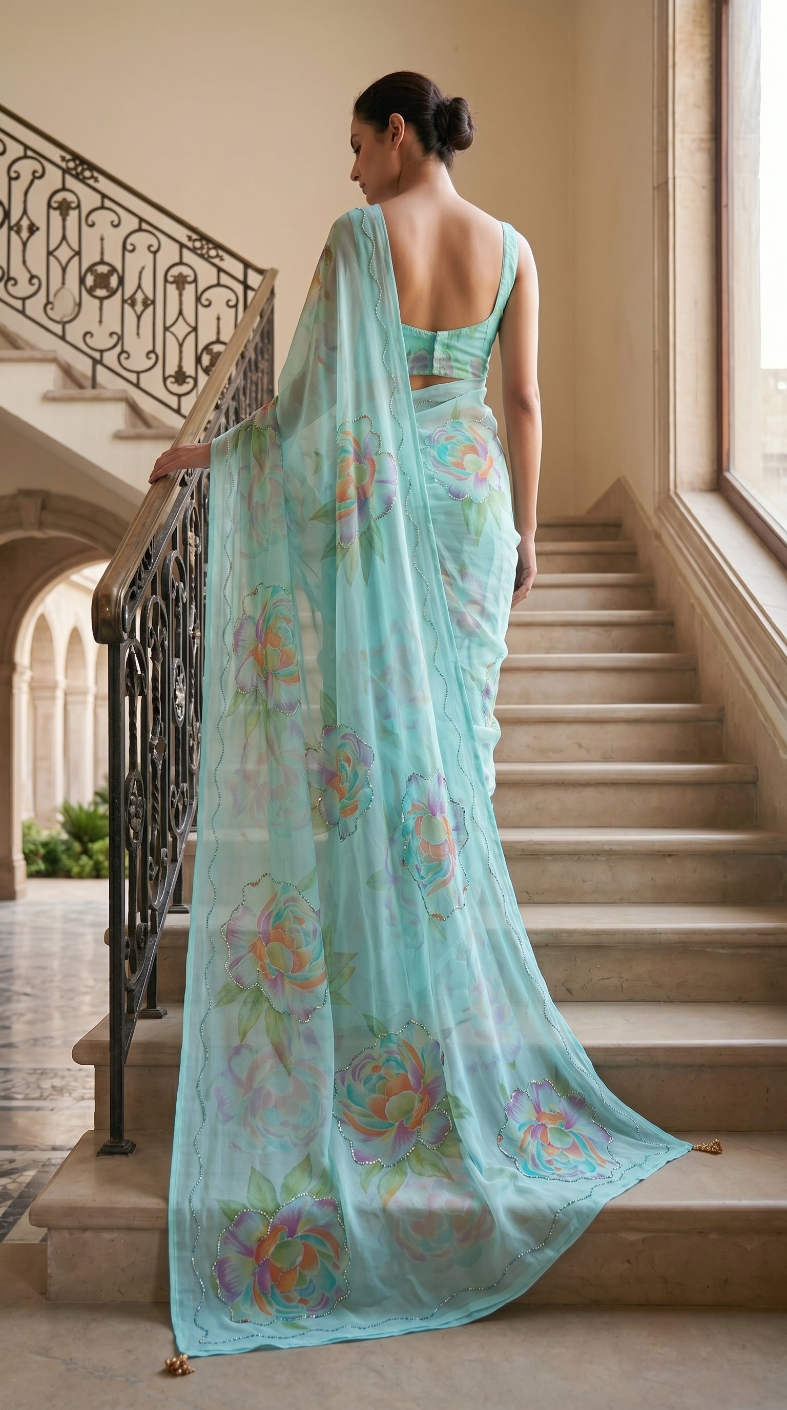 Full-length back view of a model descending marble stairs, displaying the sweeping pallu trail and scalloped edge cut work of an aqua blue floral chiffon saree