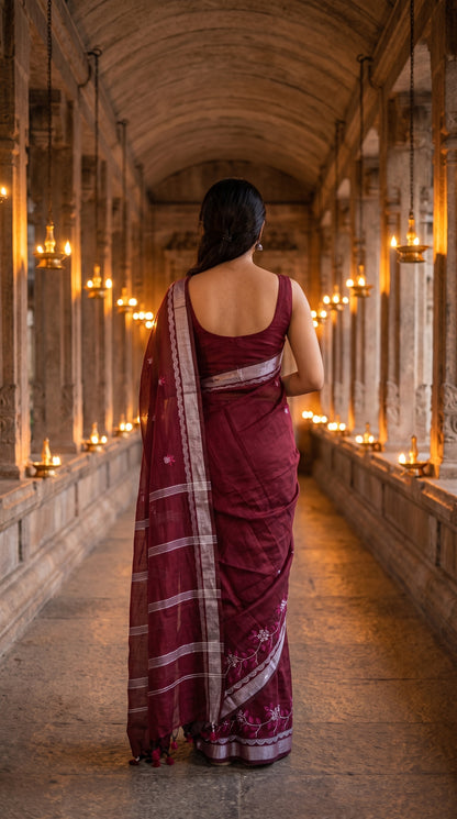 Back view of a woman walking down a long, dimly lit historic temple stone corridor lined with hanging lamps, highlighting the fluid fall and silver striped pallu of a maroon embroidered cotton linen saree