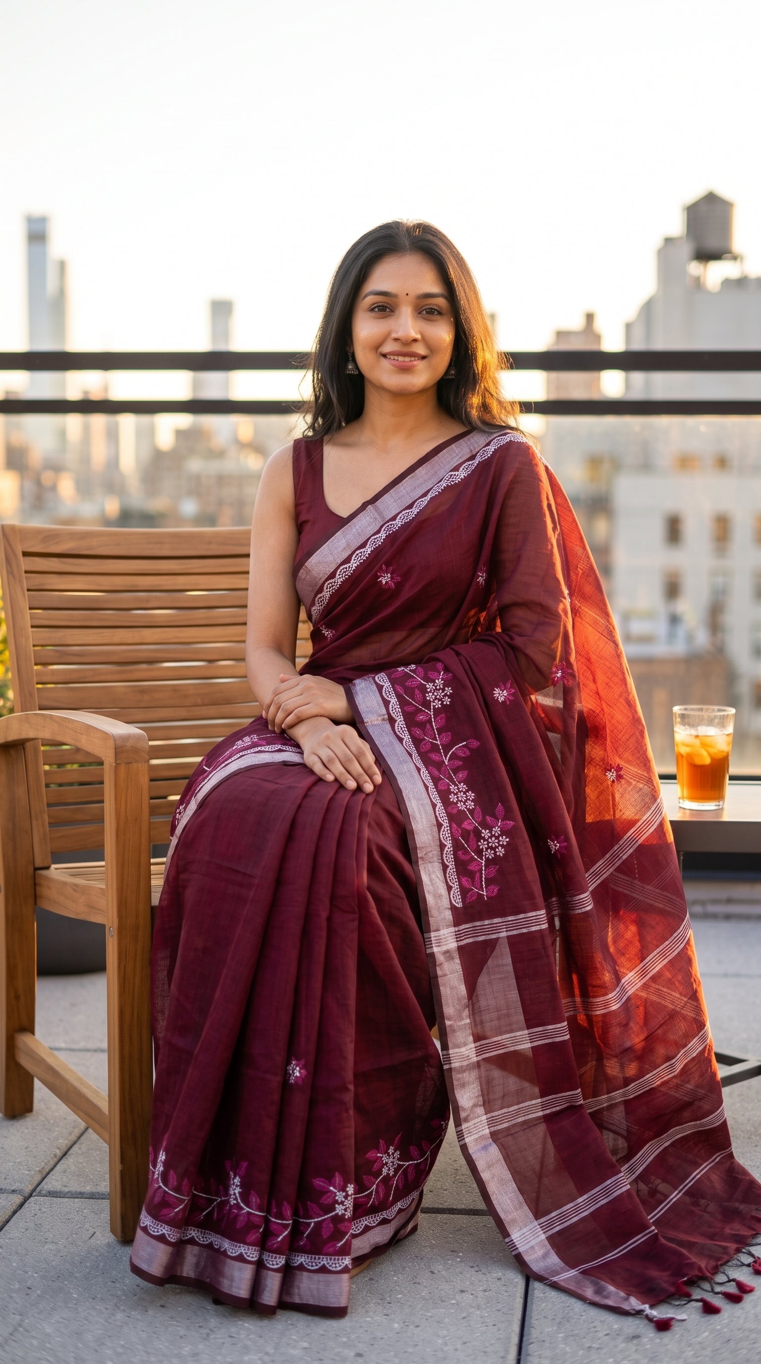 Model seated comfortably on a wooden chair on a rooftop terrace during sunset, showcasing the soft texture, neat pleats, and silver Zari highlights of a maroon cotton linen saree